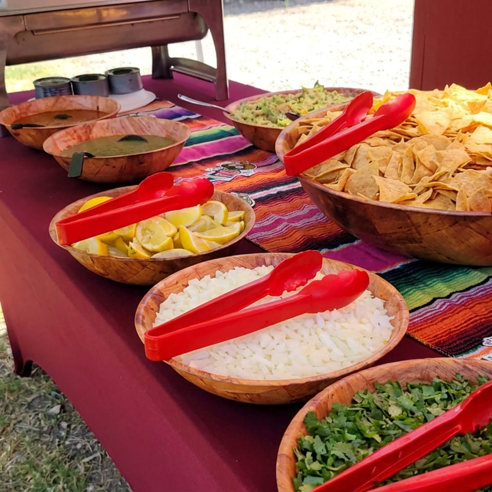 Outdoor buffet table with tortilla chips, rice, cilantro, lime wedges, salsa, guacamole, red serving tongs, and stacked plates.