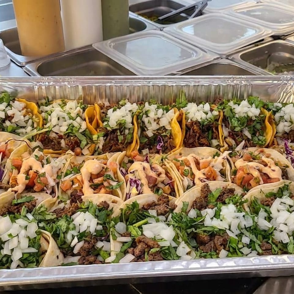 Aluminum catering tray filled with assorted tacos topped with seasoned meat, chopped onions, cilantro, vegetables, and drizzled sauce.