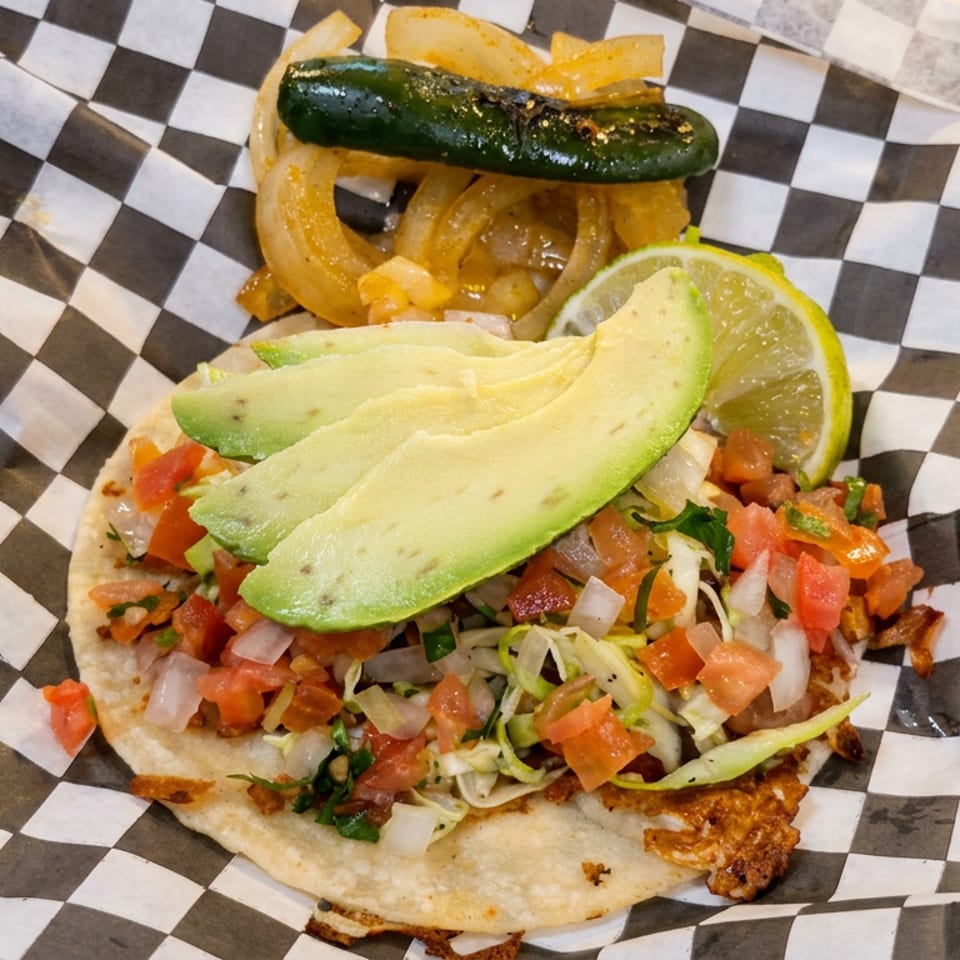 Open-faced taco topped with avocado, pico de gallo, shredded vegetables, grilled jalapeño, onions, fries, and lime.