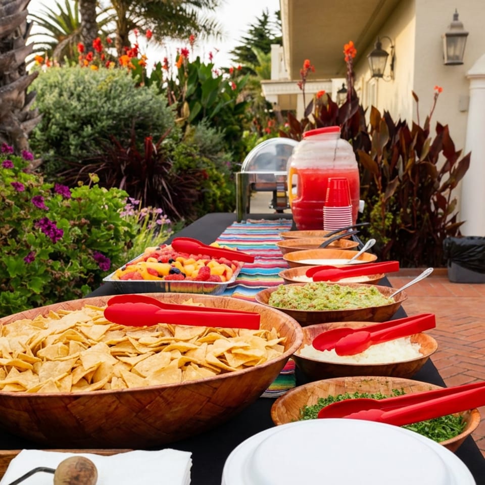 Outdoor buffet table with tortilla chips, salsa, guacamole, rice, fruit, red serving tongs, and colorful tropical garden surroundings.