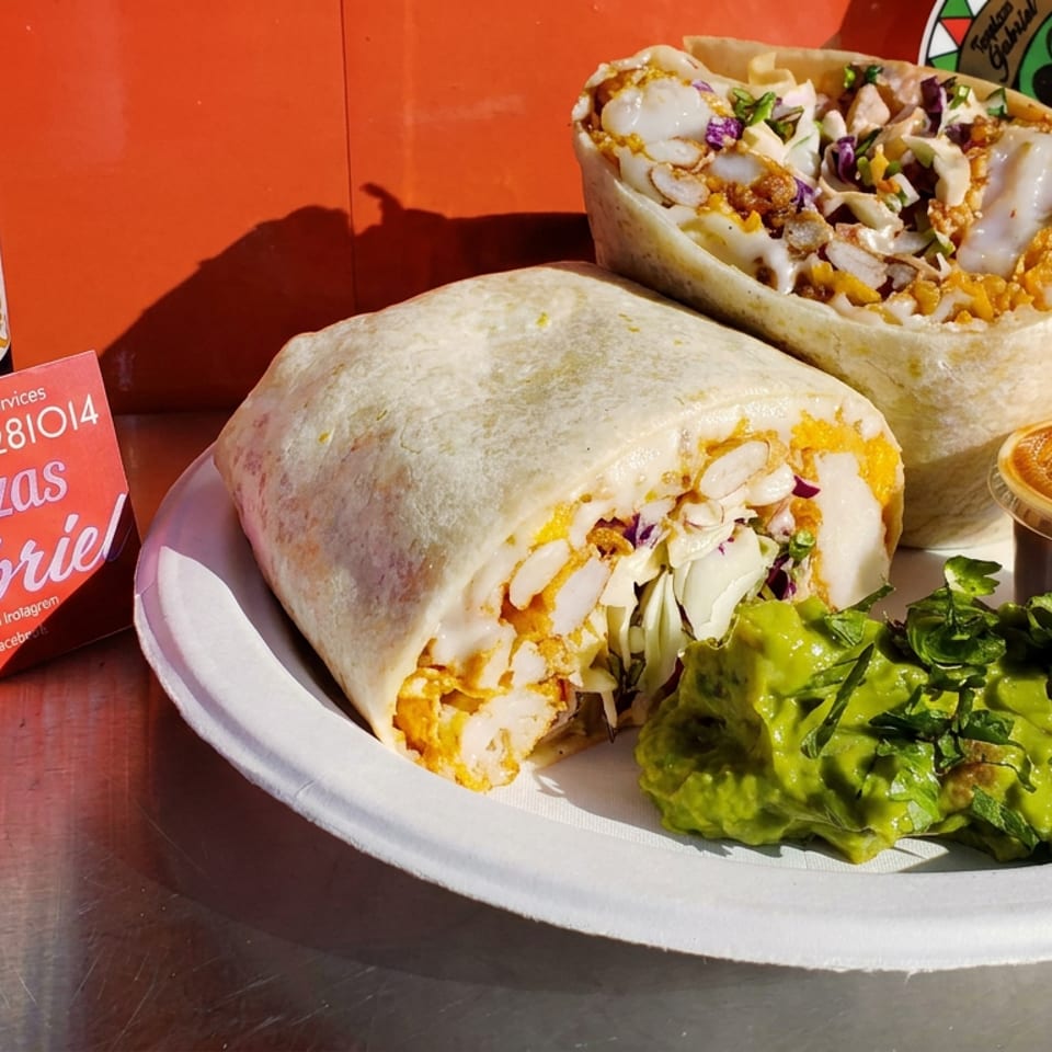 Plate of stuffed burritos with melted cheese, guacamole, and dipping sauce beside Modelo Especial beer and Taquizas Gabriela signage.
